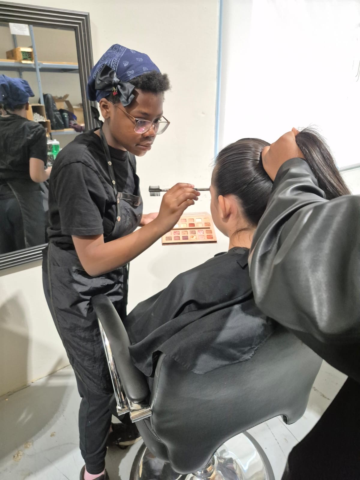 A camper applies eyeshadow to a model backstage while another holds the model's hair