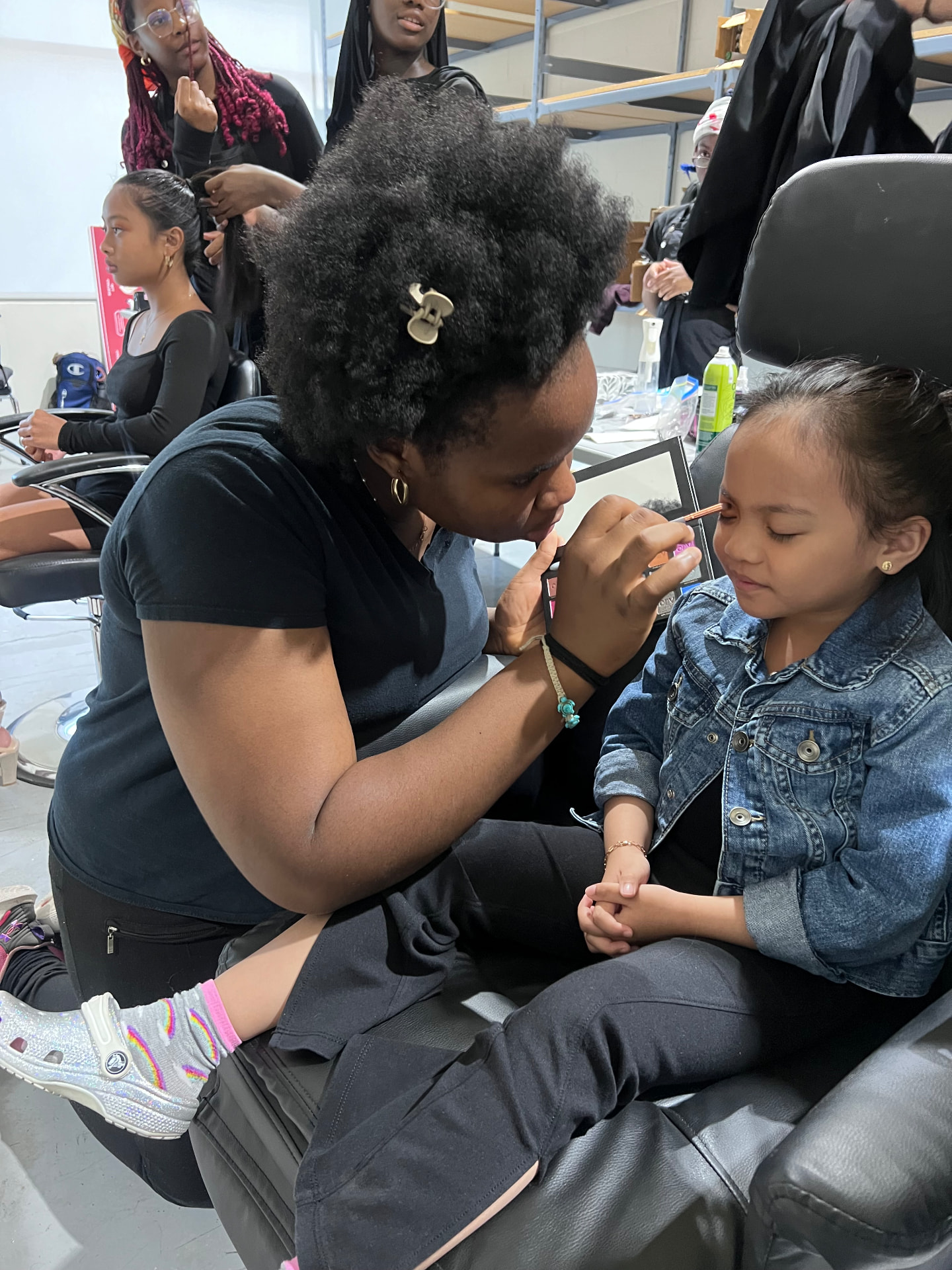 A camper applies makeup to a young client backstage at the fashion show